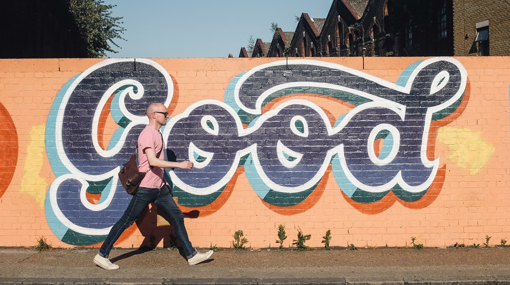 Mann mit rosa Shirt geht vor einer rosa Mauer mit dem aufgemalten Schriftzug "Good" von links nach rechts